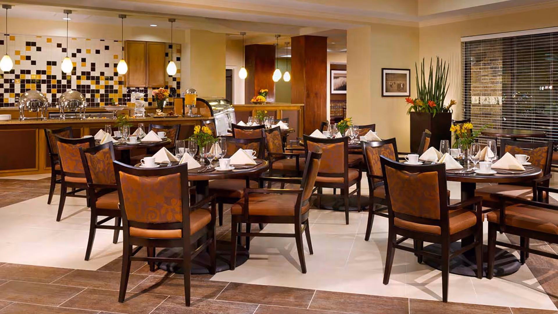 A dining room with several round tables set with white napkins, cups, glasses, and floral centerpieces. The room features a tiled floor, a counter with buffet warmers, beverage dispensers, and a decorative tiled backsplash. Pendant lights hang from the ceiling, and there is a large window with blinds and a tall plant in the corner.