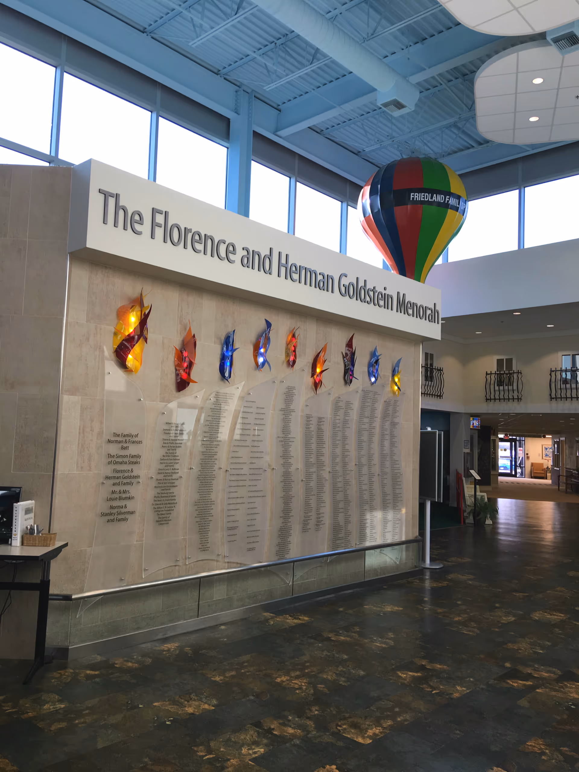 Interior lobby with a large donor wall titled "The Florence and Herman Goldstein Menorah," colorful glass sculptures, tall windows, and a hanging hot-air-balloon decoration.