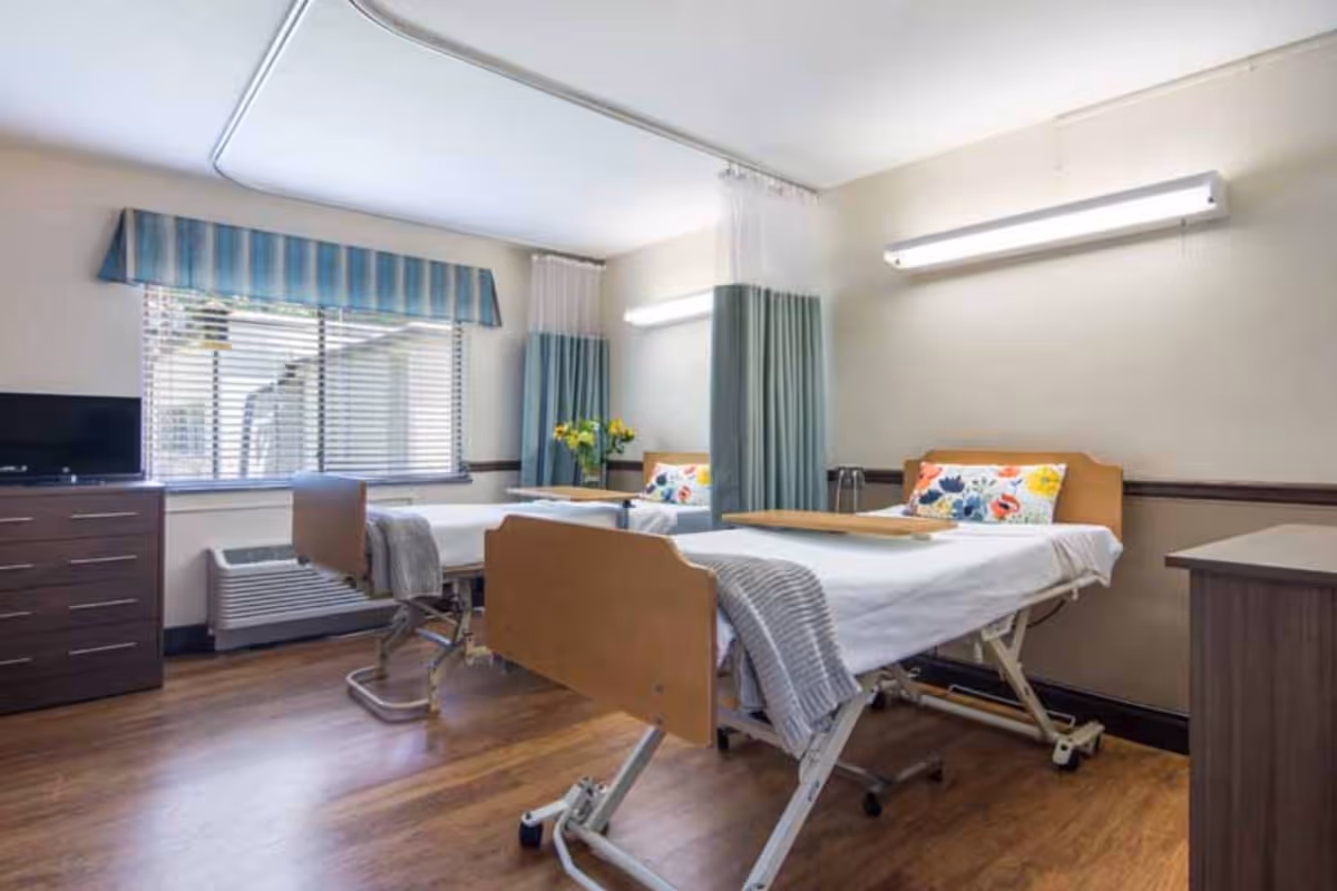 A bright and clean room in a health center with two hospital beds, each with white sheets, a colorful floral pillow, and a gray blanket. The room has wooden flooring, a window with blinds and a blue striped valance, a small TV on a dresser, and a vase with yellow flowers on a table between the beds. There are privacy curtains partially drawn around the beds and fluorescent lights on the walls.