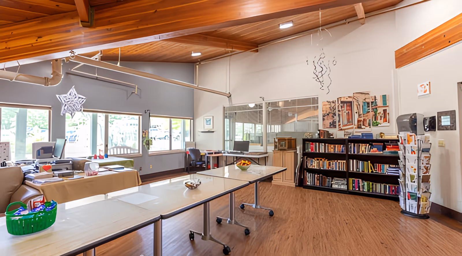 Interior of a common area in a senior living facility with long tables on wheels, a bookshelf filled with books, a rotating rack with brochures, large windows letting in natural light, and a wooden ceiling with exposed beams.