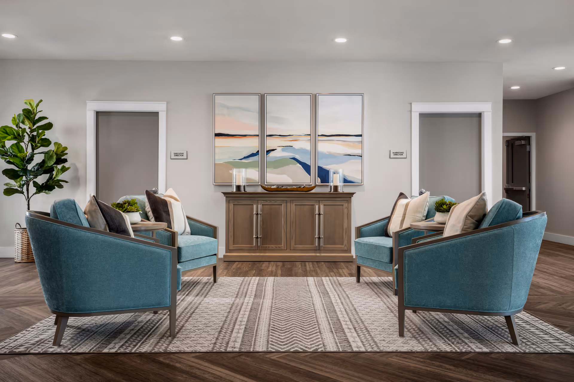 A modern and cozy seating area with four teal armchairs arranged around a patterned rug. Behind the chairs is a wooden sideboard with decorative items and a three-panel abstract landscape painting on the wall. Two doorways flank the sideboard, and a tall green plant is placed on the left side of the room. The space is well-lit with recessed ceiling lights.