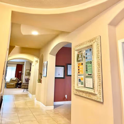 Interior hallway of a senior care home with beige walls and tiled floor. The hallway features arched doorways and a framed bulletin board with various notices on the right wall. In the background, there is a living area with chairs and a window with curtains.