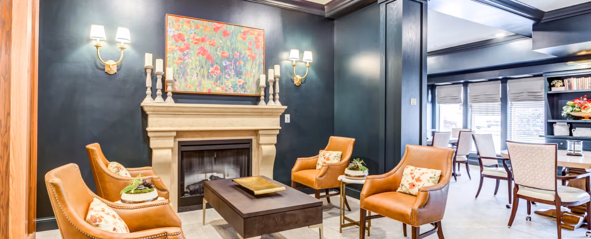 A cozy senior living common area featuring four tan leather armchairs with floral cushions arranged around a dark wooden coffee table. Behind the chairs is a beige stone fireplace adorned with multiple white candles and a colorful floral painting above it. The walls are painted dark blue, and there are two wall sconces with white lampshades on either side of the fireplace. In the background, there is a dining area with several chairs and tables near large windows with blinds.