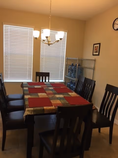 Dining room with a rectangular table covered by a multicolored tablecloth and six dark wooden chairs. Six red placemats are arranged on the table. Two windows with closed blinds are in the background, along with a metal shelving unit holding water bottles. A ceiling light fixture with five bulbs hangs above the table, and a clock and framed picture are on the beige walls.