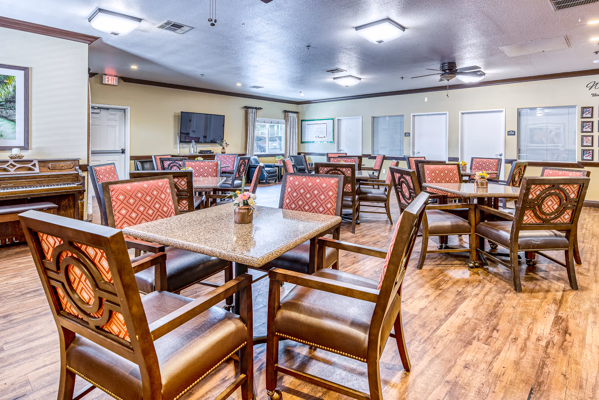 A spacious dining room with multiple square and round tables surrounded by wooden chairs with red patterned cushions. The room has wood flooring, a piano on the left side, a wall-mounted TV, and windows with curtains. The walls are light-colored with dark trim, and there are ceiling fans and recessed lighting.