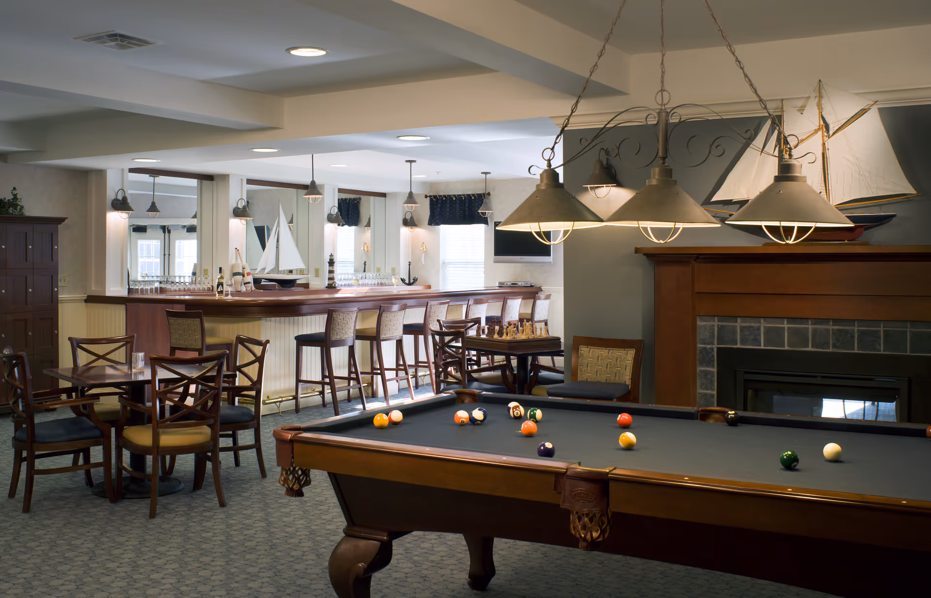 A cozy recreational room featuring a pool table with billiard balls scattered on it, a chessboard on a small table, several wooden chairs and tables, a bar area with high stools, decorative model sailboats, and a fireplace with a wooden mantle.