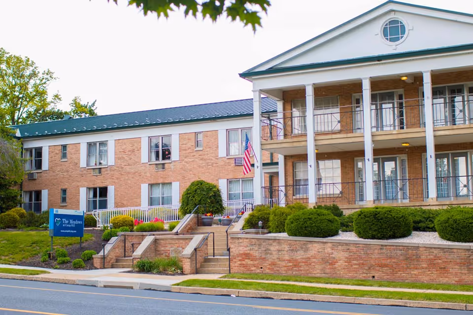 Exterior view of a two-story brick senior living facility with white columns and balconies. There are well-maintained bushes and landscaping in front, a set of stairs leading up to the entrance, and an American flag displayed near the door. A blue sign near the sidewalk reads 'The Meadows at Camp Hill.'