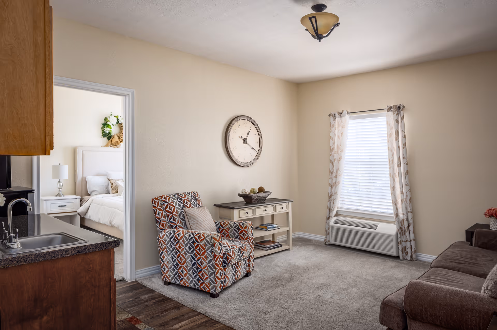 A cozy living room with a patterned armchair, a small console table with decorative items, a large wall clock, a window with floral curtains and a wall-mounted air conditioning unit below it. To the left, part of a kitchen countertop and sink are visible, and through an open doorway, a bedroom with a bed, nightstand, lamp, and floral wreath on the wall can be seen.