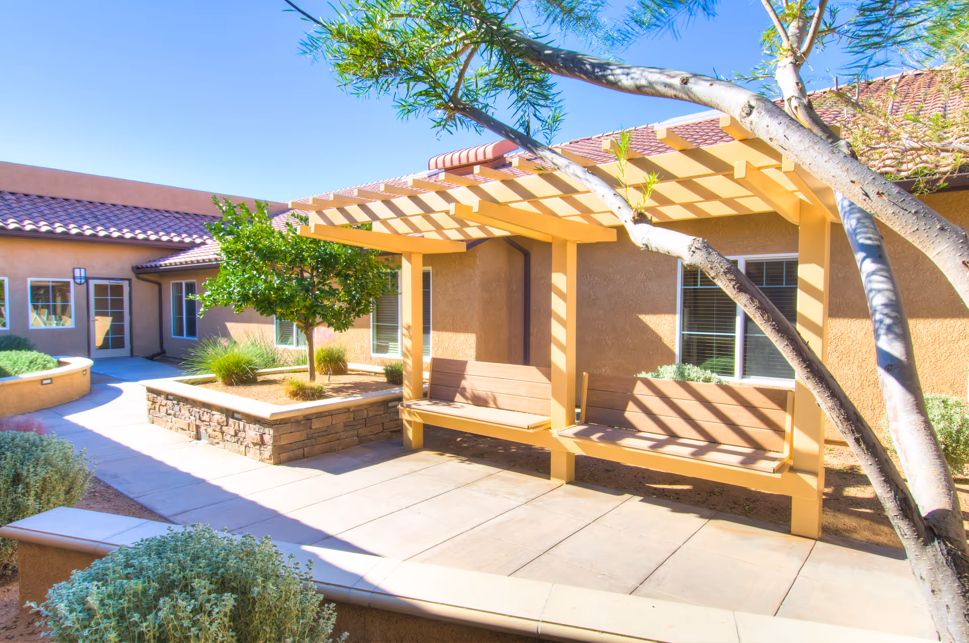 Outdoor courtyard area at Lake View Terrace Memory Care Residence featuring a shaded wooden bench swing under a pergola, surrounded by desert landscaping with small trees and shrubs, and a building with tan stucco walls and red tile roof in the background.