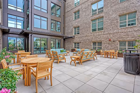 Sunlit courtyard patio with wooden tables, lounge chairs, and planters surrounded by a multi-story brick building.