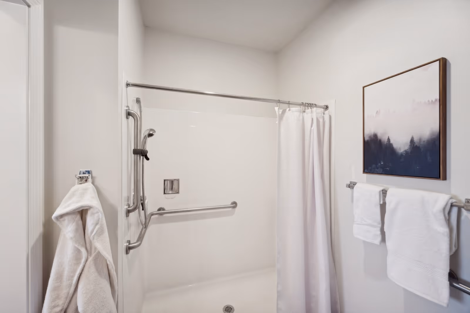 A bathroom shower area with white walls and a white shower curtain. The shower has metal grab bars and a handheld showerhead. Two white towels hang on a towel rack on the right wall, below a framed picture of a misty forest. A white bathrobe hangs on a hook on the left wall.