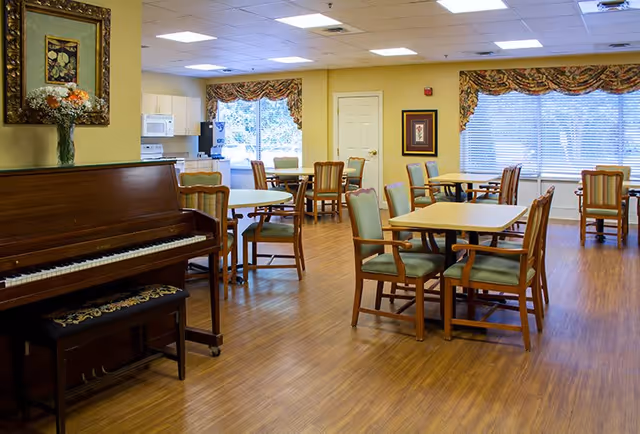 A well-lit dining room with several wooden tables and chairs arranged neatly on a wooden floor. A piano with a floral cushioned bench is positioned on the left side of the room. The walls are painted light yellow, decorated with framed artwork and a vase of flowers on the piano. Large windows with patterned valances allow natural light to fill the space.
