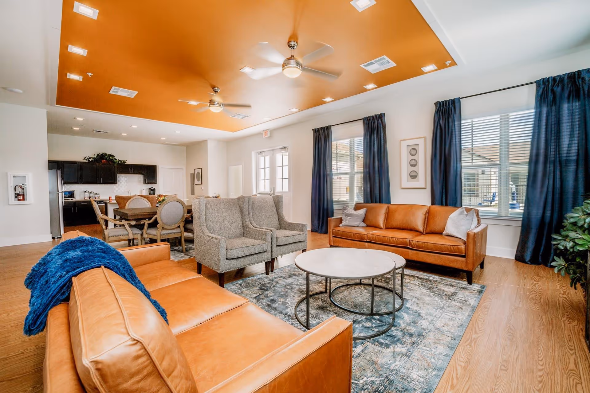 Bright common living area with tan leather sofas, gray armchairs, a round coffee table, dining area and kitchen in the background.