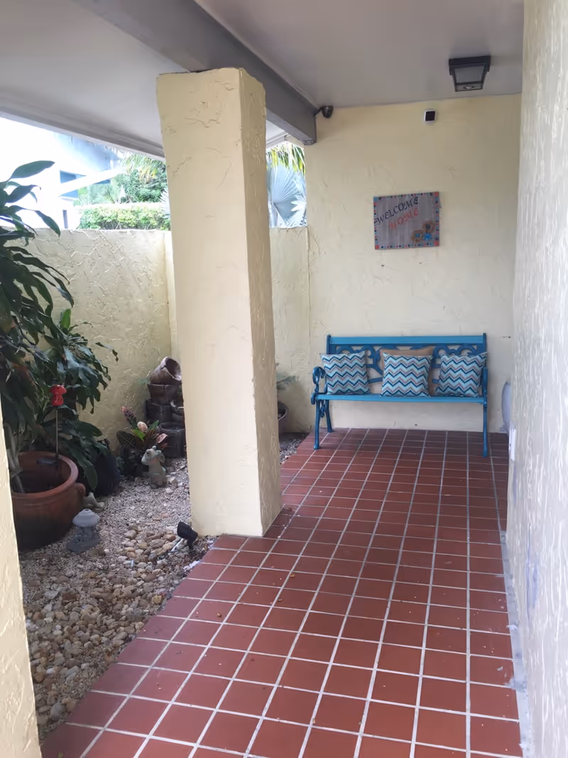 Covered exterior entryway with red tiled floor, a blue bench with patterned pillows, and potted plants along a pebble garden.