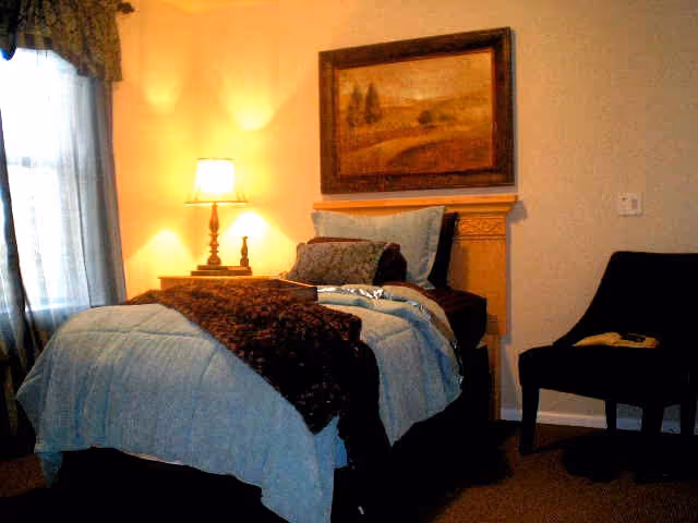 A cozy bedroom with a single bed covered in light blue bedding and a dark brown throw blanket. There is a wooden headboard and a framed landscape painting above it. A bedside table with a lamp is next to the bed, and a dark upholstered chair with a book on it is positioned to the right. Light filters in through a window with sheer curtains and a valance.
