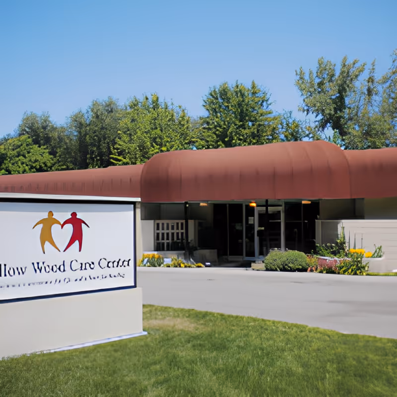 Front entrance of Willow Wood Care Center with a large maroon awning, landscaped beds, and a sign on the lawn.