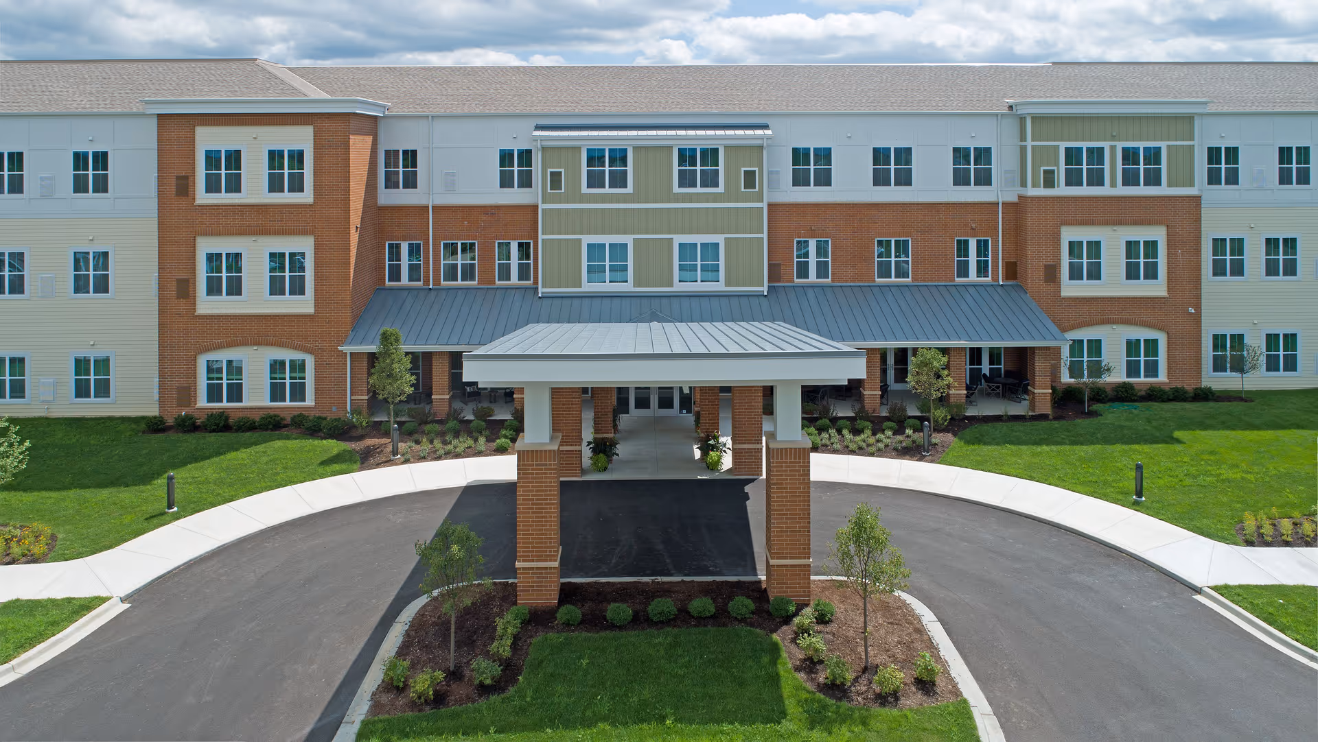 Front entrance and porte-cochère of a three-story assisted living building with a circular driveway and landscaped lawn.