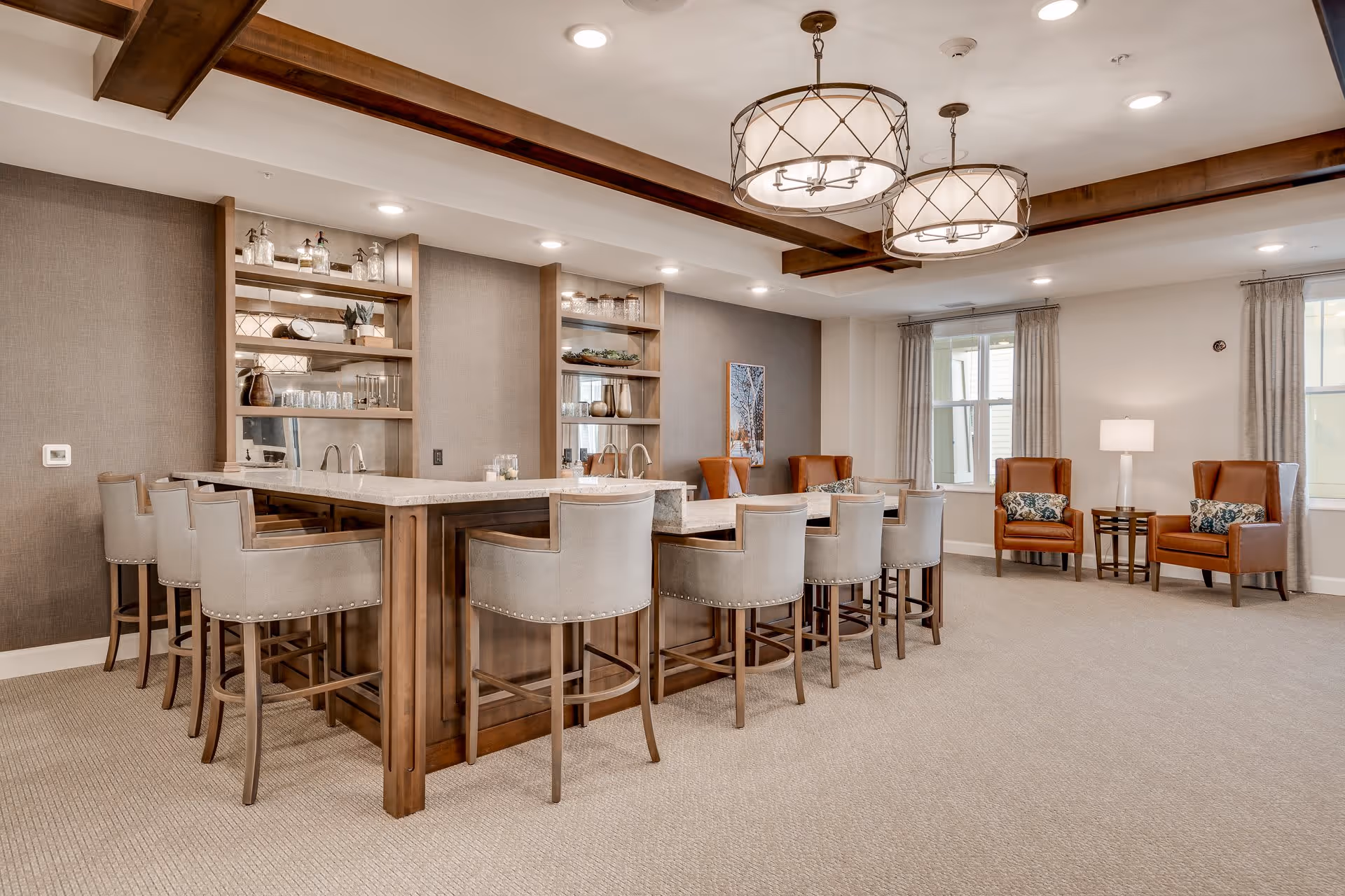A spacious and well-lit common area with a large wooden bar counter surrounded by cushioned bar stools. The bar has shelves with glassware and decorative items. In the background, there are two large windows with curtains, two brown leather armchairs with patterned cushions, and a small side table with a lamp. The ceiling features wooden beams and two large pendant lights.