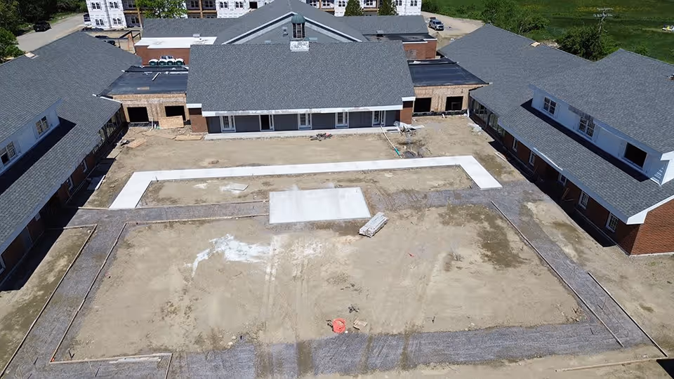 Aerial view of a senior living facility named Wiscasset Green under construction, showing multiple buildings with gray roofs surrounding a large dirt courtyard with newly laid concrete pathways.