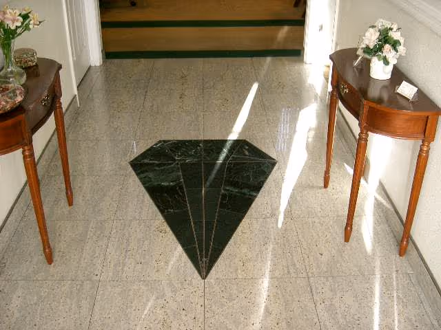 A hallway with a polished tile floor featuring a dark green diamond-shaped inlay in the center. On either side of the hallway are small wooden tables with curved legs, each holding a vase with flowers and decorative items. Sunlight is streaming in from the right side, casting shadows on the floor.