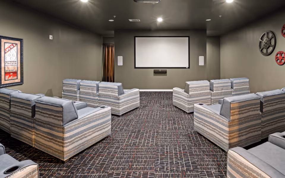 A small theater room with multiple rows of cushioned armchairs facing a large blank projection screen on the wall. The room has dark walls and ceiling with recessed lighting, patterned carpet, and decorative film reel wall art on the right side. A framed poster is visible on the left wall.