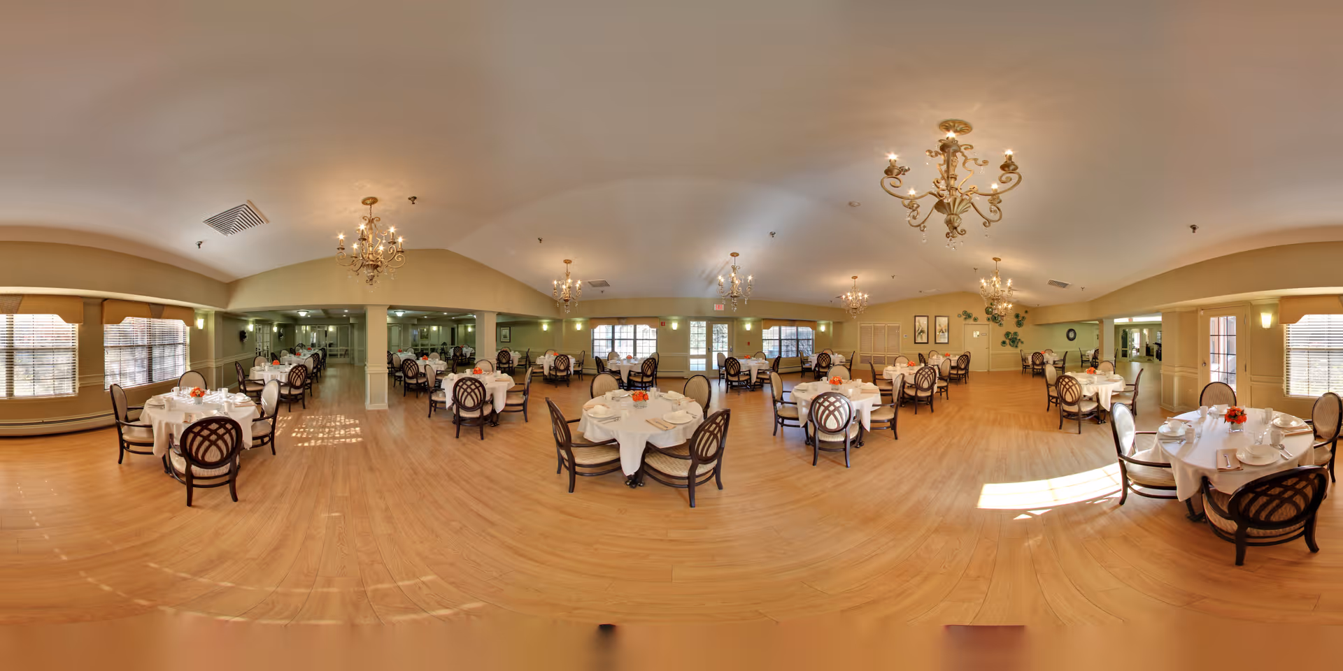 A spacious senior community dining room with multiple round tables covered in white tablecloths, each set with plates, cups, and small floral centerpieces. The room features wooden flooring, large windows with blinds allowing natural light, elegant chandeliers hanging from the ceiling, and light-colored walls adorned with framed artwork and plants.