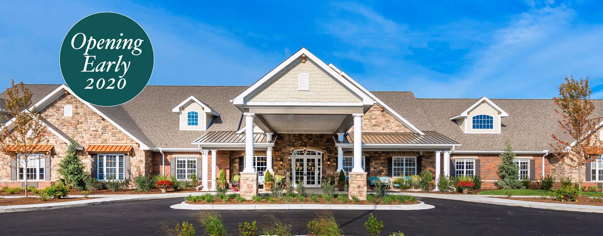 Front exterior view of a large, single-story senior living facility building with a stone and brick facade, white columns at the entrance, and a circular driveway. There are small trees and shrubs planted around the building. A green circular sign on the left side says 'Opening Early 2020'.