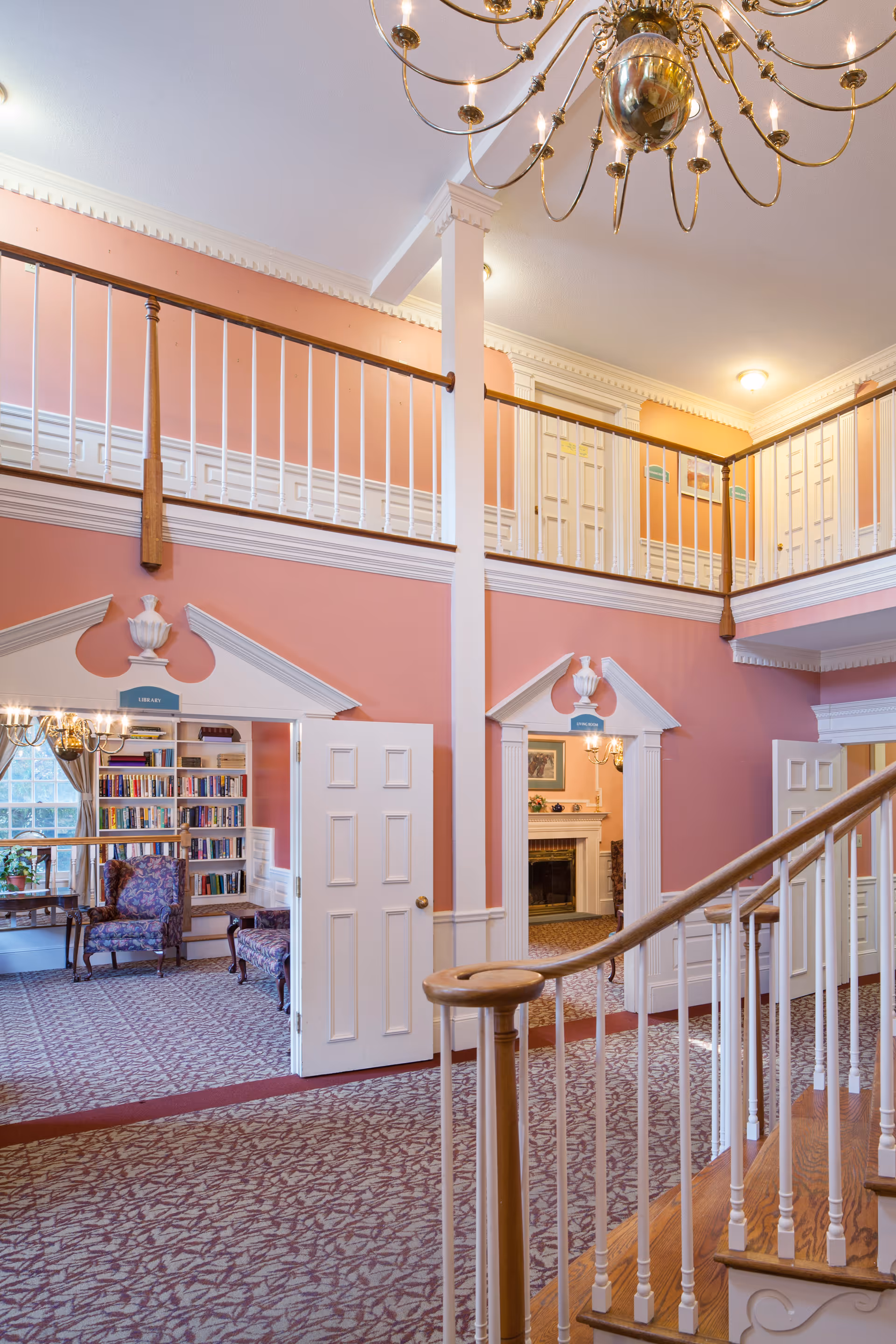 Two-story interior common area with a wooden staircase, upstairs balcony, seating, bookshelves, and a fireplace.