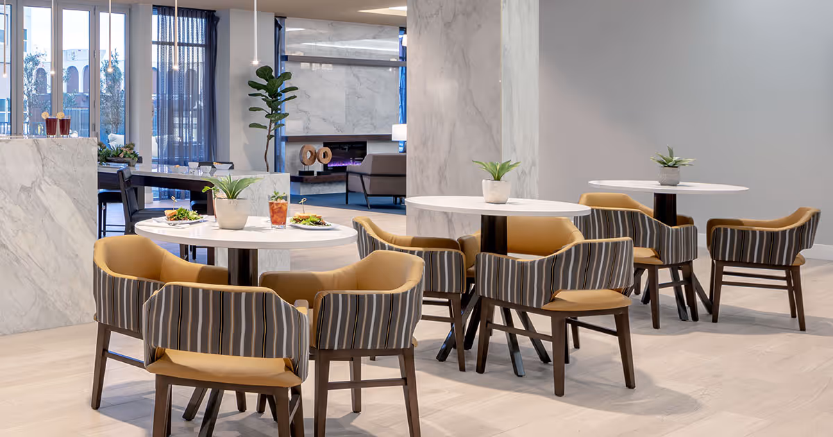 Modern communal dining area with round white tables, striped cushioned chairs, and small potted plants.