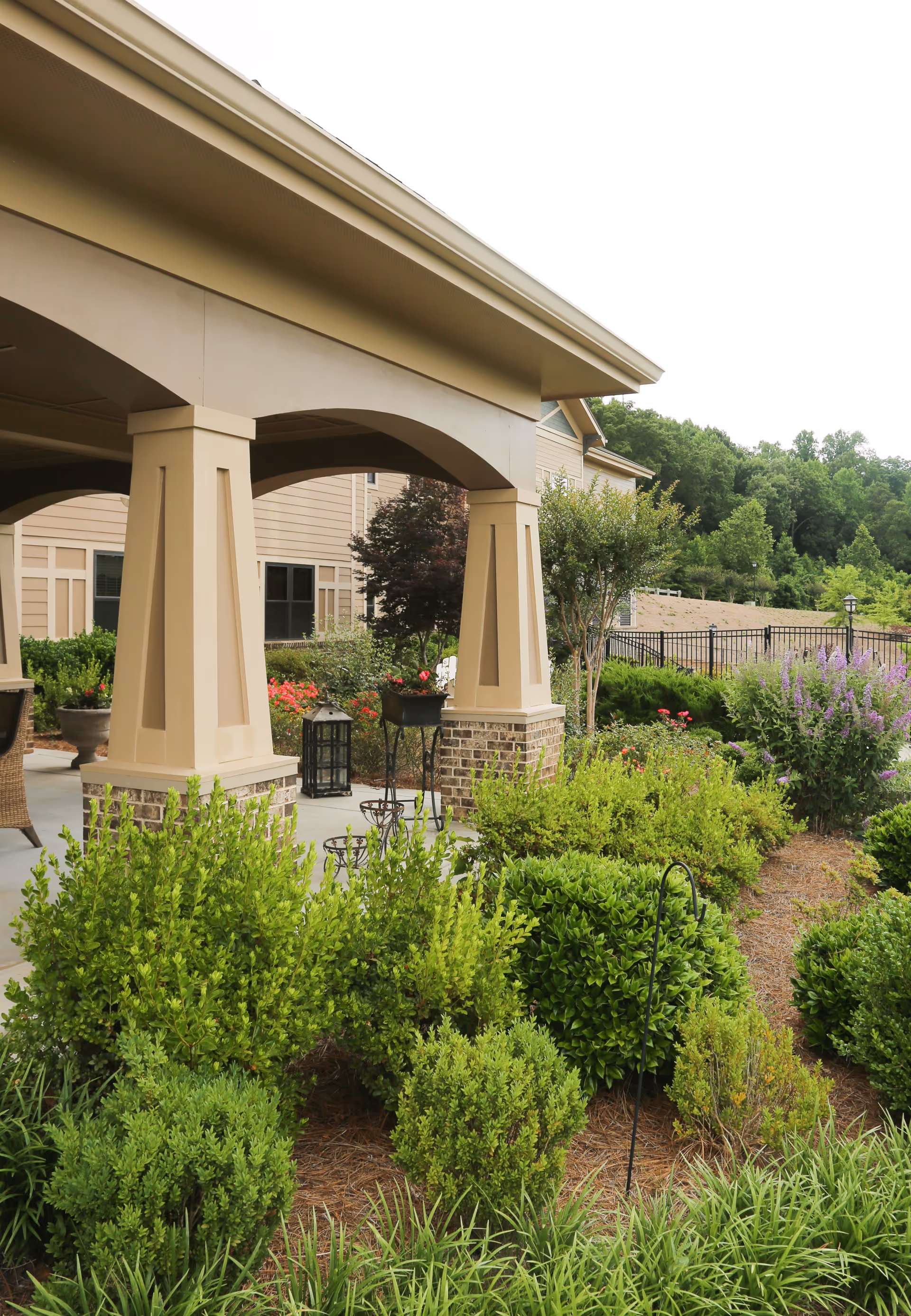 Covered outdoor patio with large columns and surrounding landscaped shrubs and flowers at the front of a senior living facility.