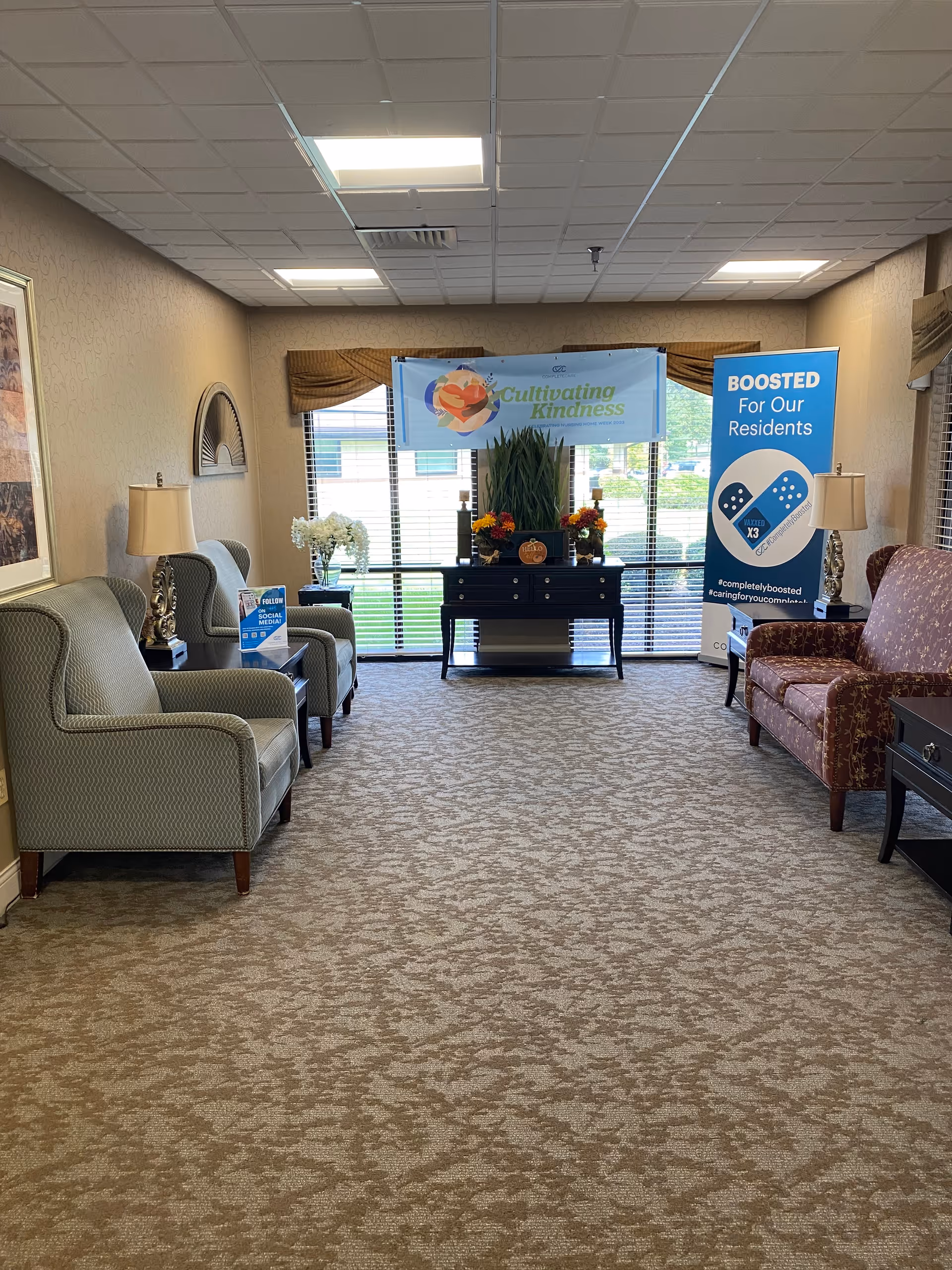 A well-lit sitting area in a senior living facility with patterned carpet, two armchairs on the left, a patterned sofa on the right, and a black console table with plants and flowers in front of large windows. Two banners are displayed behind the table, one reading 'Cultivating Kindness' and the other 'BOOSTED For Our Residents' with an image of band-aids.