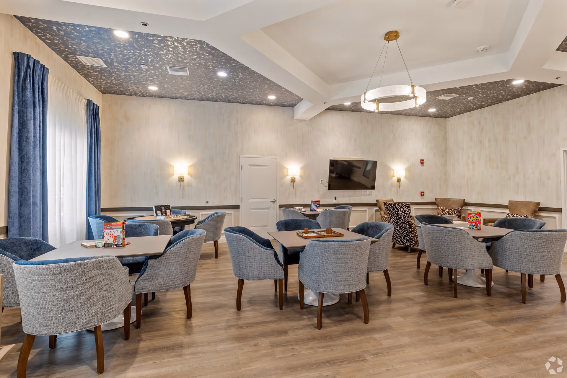 A well-lit common room with multiple tables and comfortable chairs arranged for group seating. The room features light-colored walls, a patterned ceiling, wall sconces, a flat-screen TV mounted on the wall, and large windows with blue curtains allowing natural light to enter. Some tables have board games and card games placed on them.