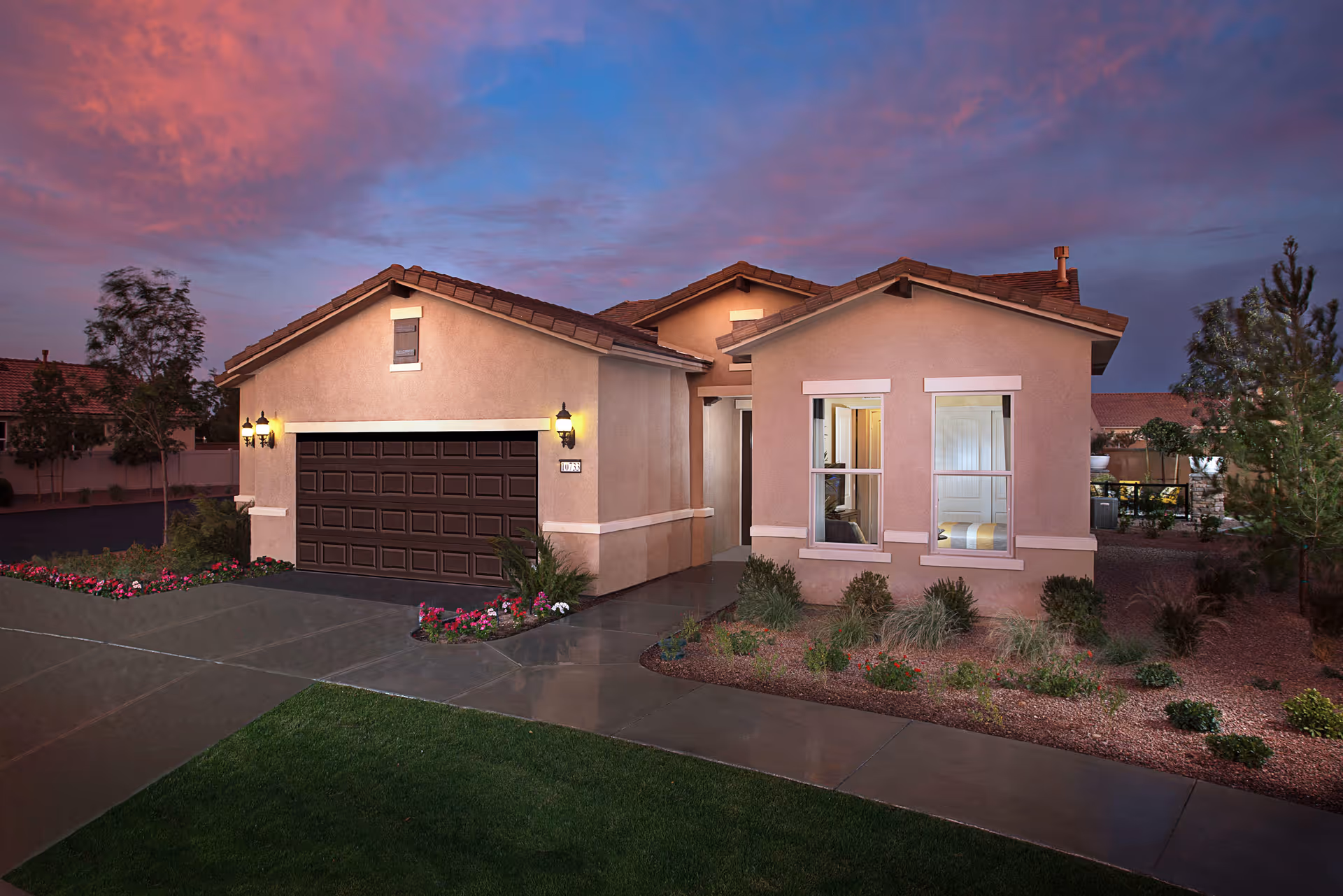 Exterior view of a single-story house at dusk with a two-car garage, illuminated wall lanterns, a well-maintained front yard with grass, flowers, and shrubs, and a colorful sky with pink and purple clouds.