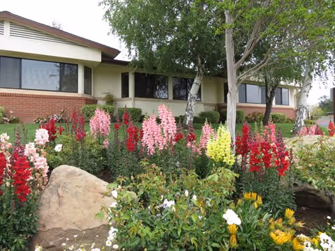 Exterior view of a single-story brick and stucco building with large windows, surrounded by a garden featuring colorful flowers including pink, red, yellow, and white blooms, along with green shrubs and trees.