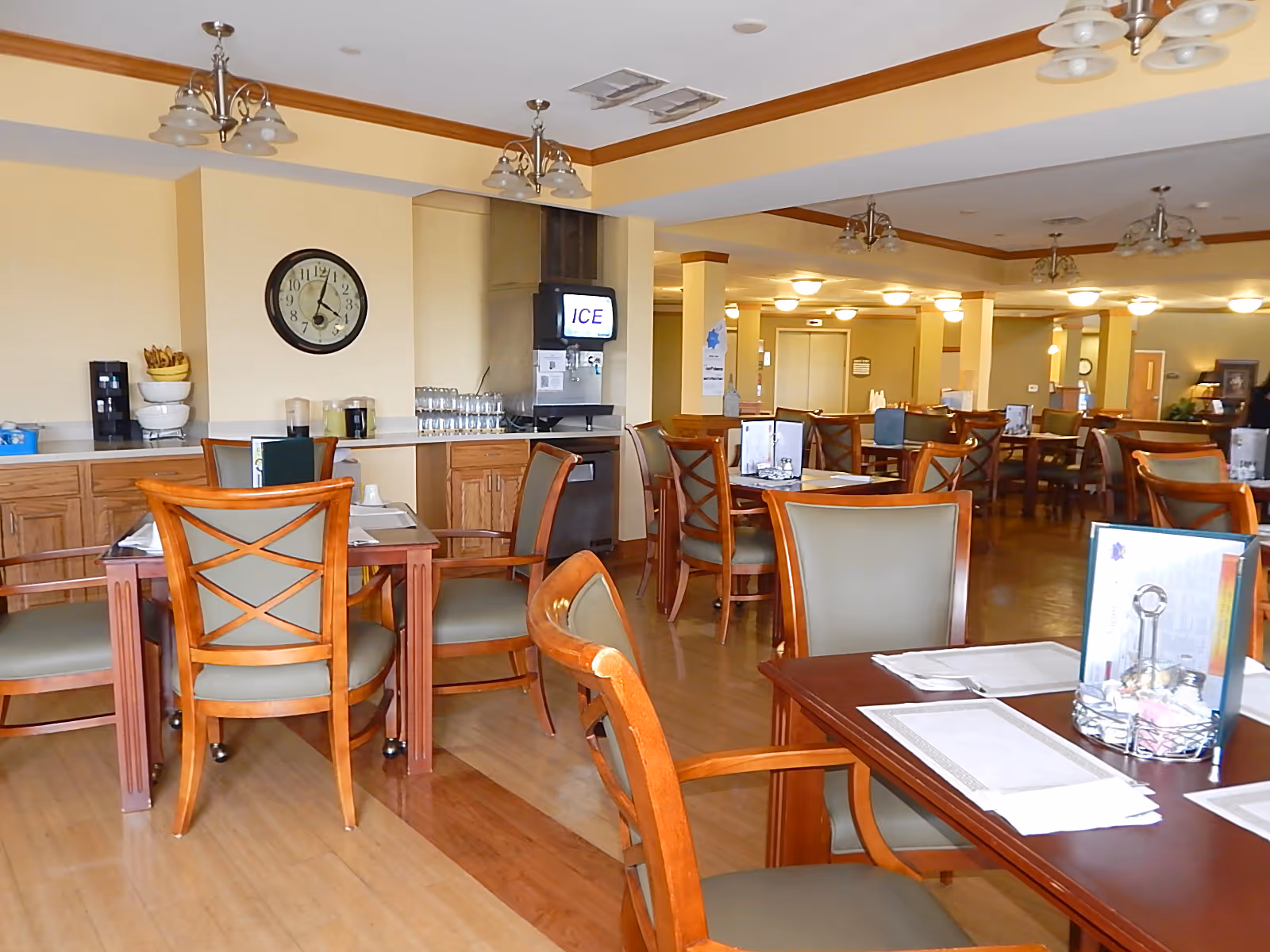 A spacious dining room with multiple wooden tables and chairs arranged neatly. The room has a light yellow wall color with wooden trim and ceiling light fixtures. There is a clock on the wall, an ice dispenser, and a counter with glasses and bowls. Menus and place settings are on the tables.
