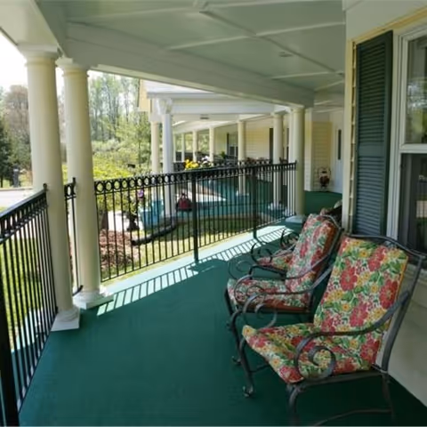Covered outdoor porch area with green carpet flooring, black metal railing, and two cushioned chairs with floral patterned upholstery. The porch overlooks a garden area with trees and shrubs in the background.