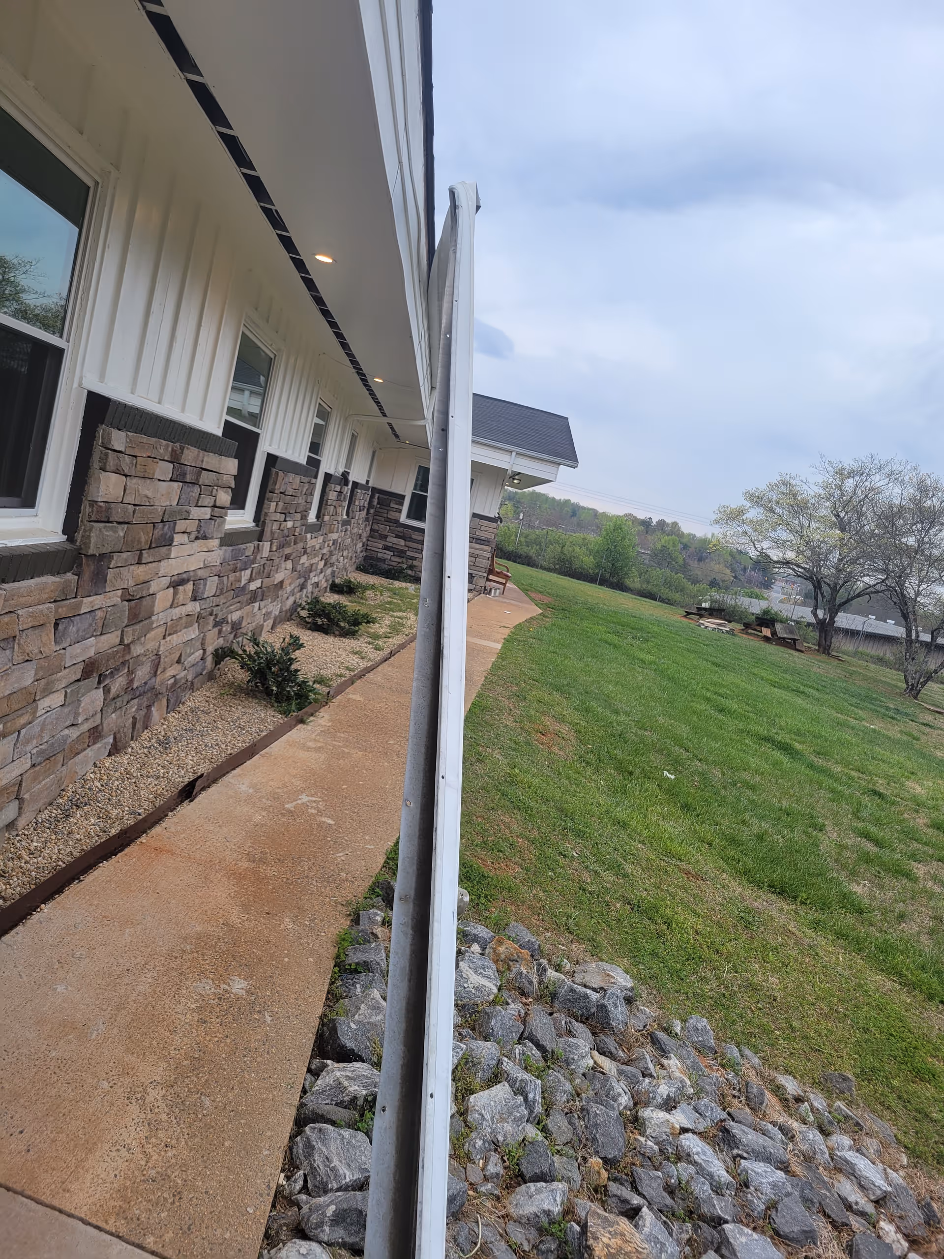 Side view of a building with stone and white siding exterior, a concrete walkway, small bushes along the building, and a grassy area with trees and rocks on the right side under a cloudy sky.