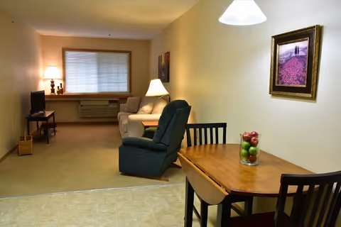 A cozy living area in a senior care facility featuring a small wooden dining table with four chairs, a glass vase with apples as a centerpiece, a green recliner chair, a beige sofa, a side table with a lamp, a television on a stand, and framed artwork on the walls. The room has a window with blinds and soft lighting.