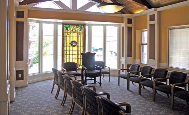 A well-lit room with a semi-circle arrangement of chairs facing a large stained glass window. The room has a high ceiling with exposed beams and a hanging light fixture. The walls are painted beige with white trim, and there are large windows letting in natural light.