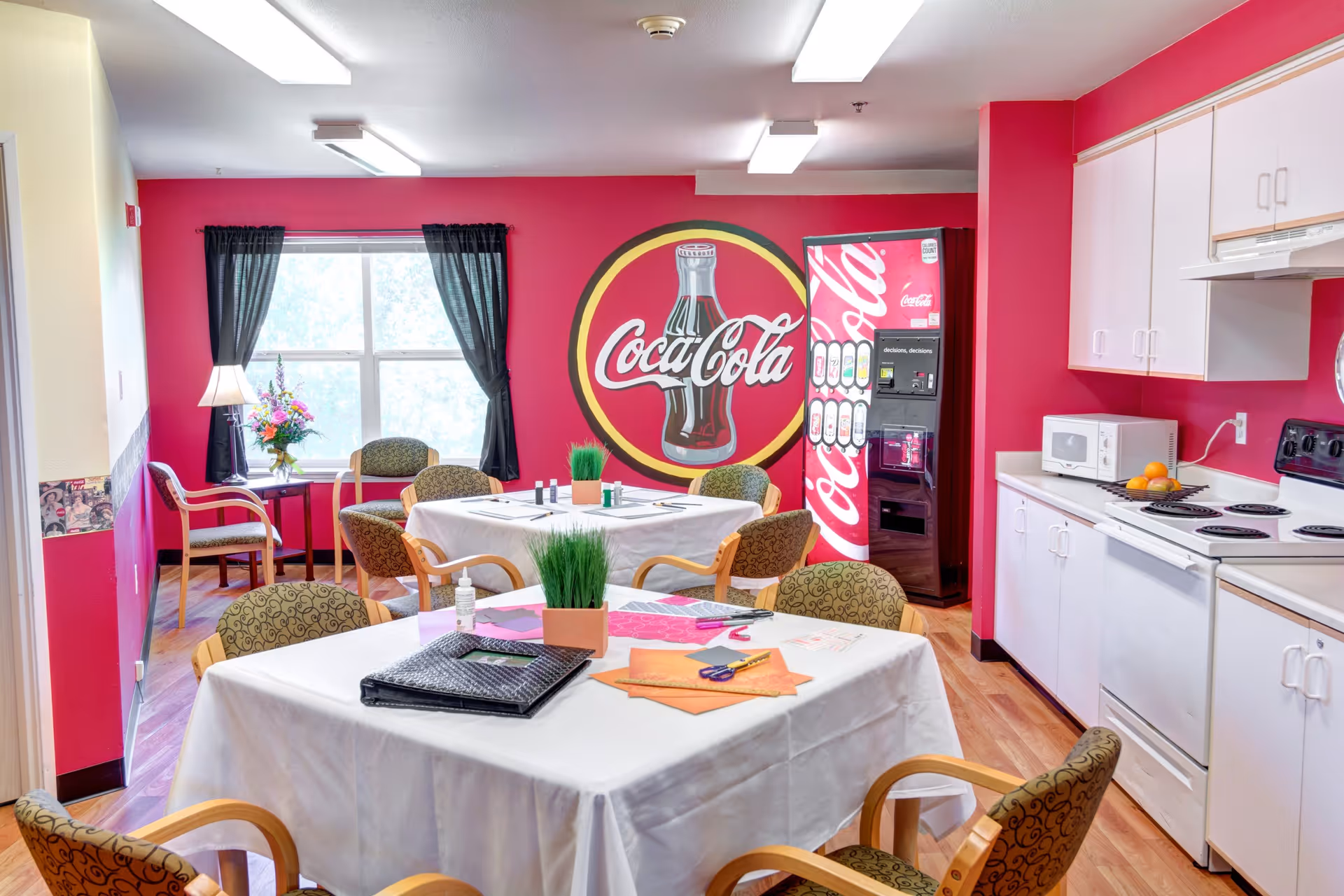 A bright room with pink walls featuring a large Coca-Cola logo and a Coca-Cola vending machine. The room has two tables covered with white tablecloths, surrounded by patterned chairs. On the tables are craft supplies, a small potted plant, and a bottle of hand sanitizer. The room also includes a kitchen area with white cabinets, a microwave, stove, and a bowl of oranges. A window with black curtains and a small side table with a lamp and flowers are visible in the background.