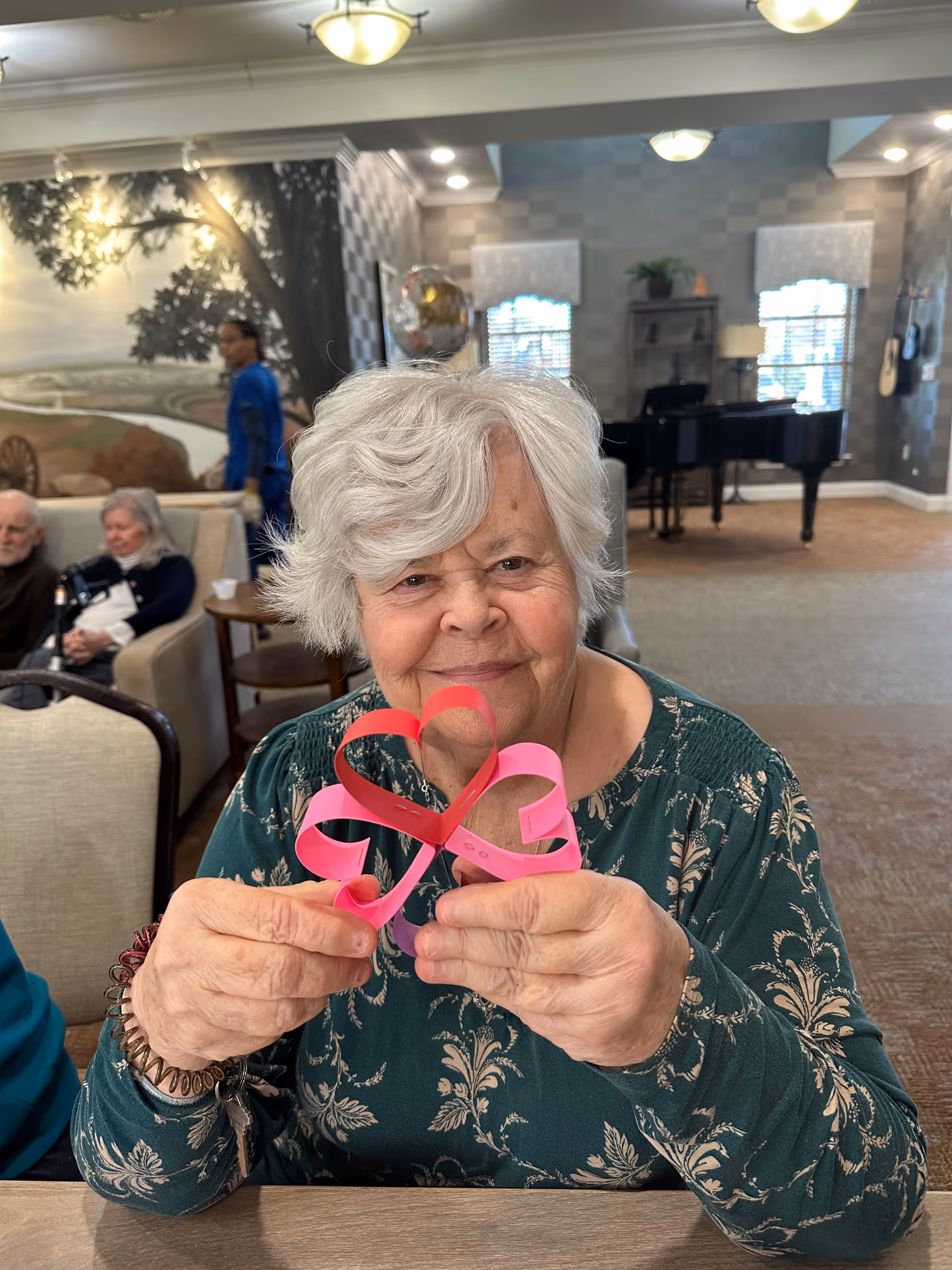 An elderly woman with white hair sitting at a table in a common area, holding a pink paper craft shaped like a heart. In the background, there are other elderly people seated on couches, a person walking, a mural on the wall, and a black grand piano near windows with blinds.