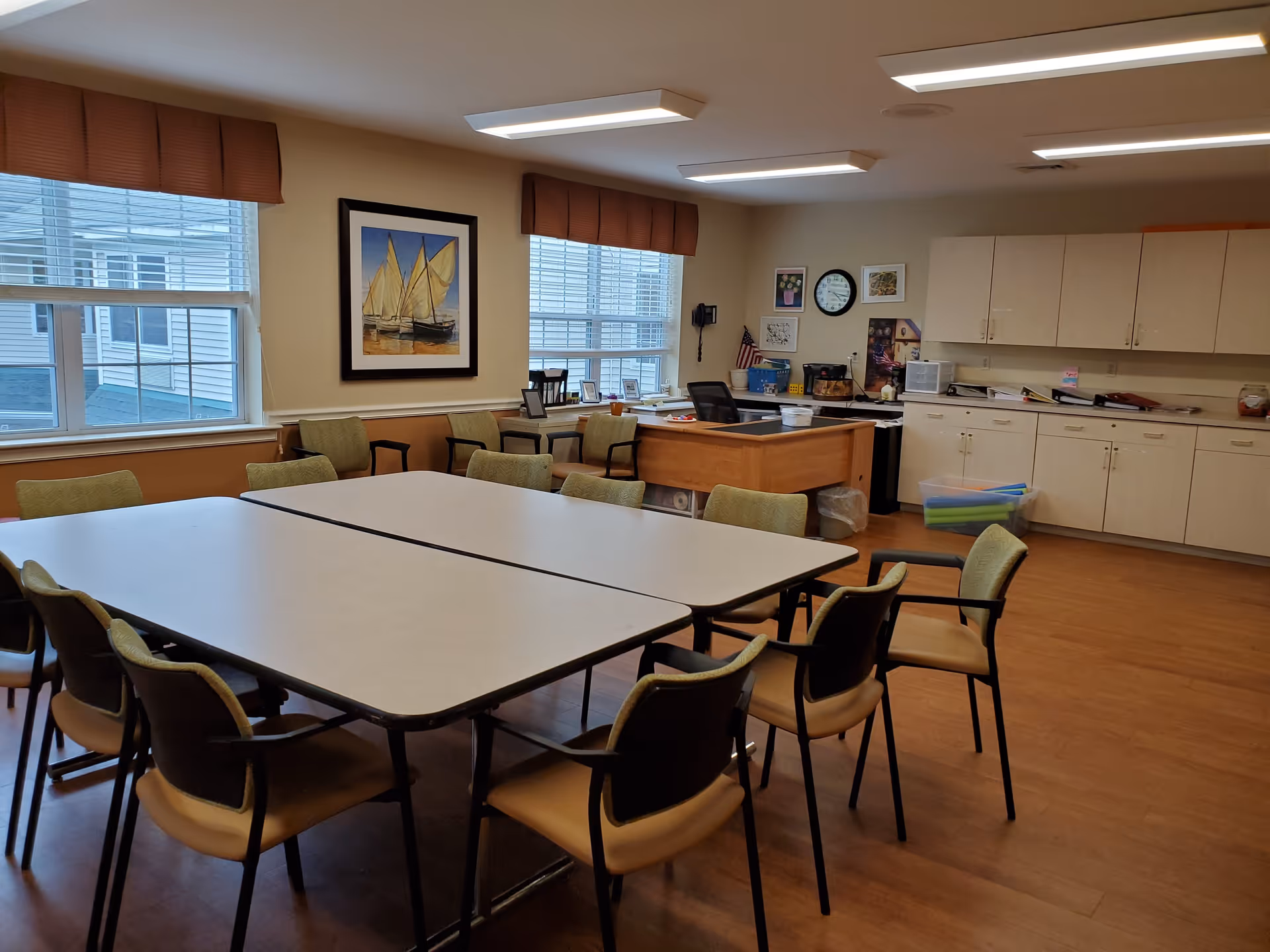 A well-lit room with two rectangular tables pushed together surrounded by chairs with green cushions. The room has large windows with brown valances, a framed painting of sailboats on the wall, a desk with office supplies, and white cabinets along one wall. The floor is wooden, and the ceiling has rectangular fluorescent lights.