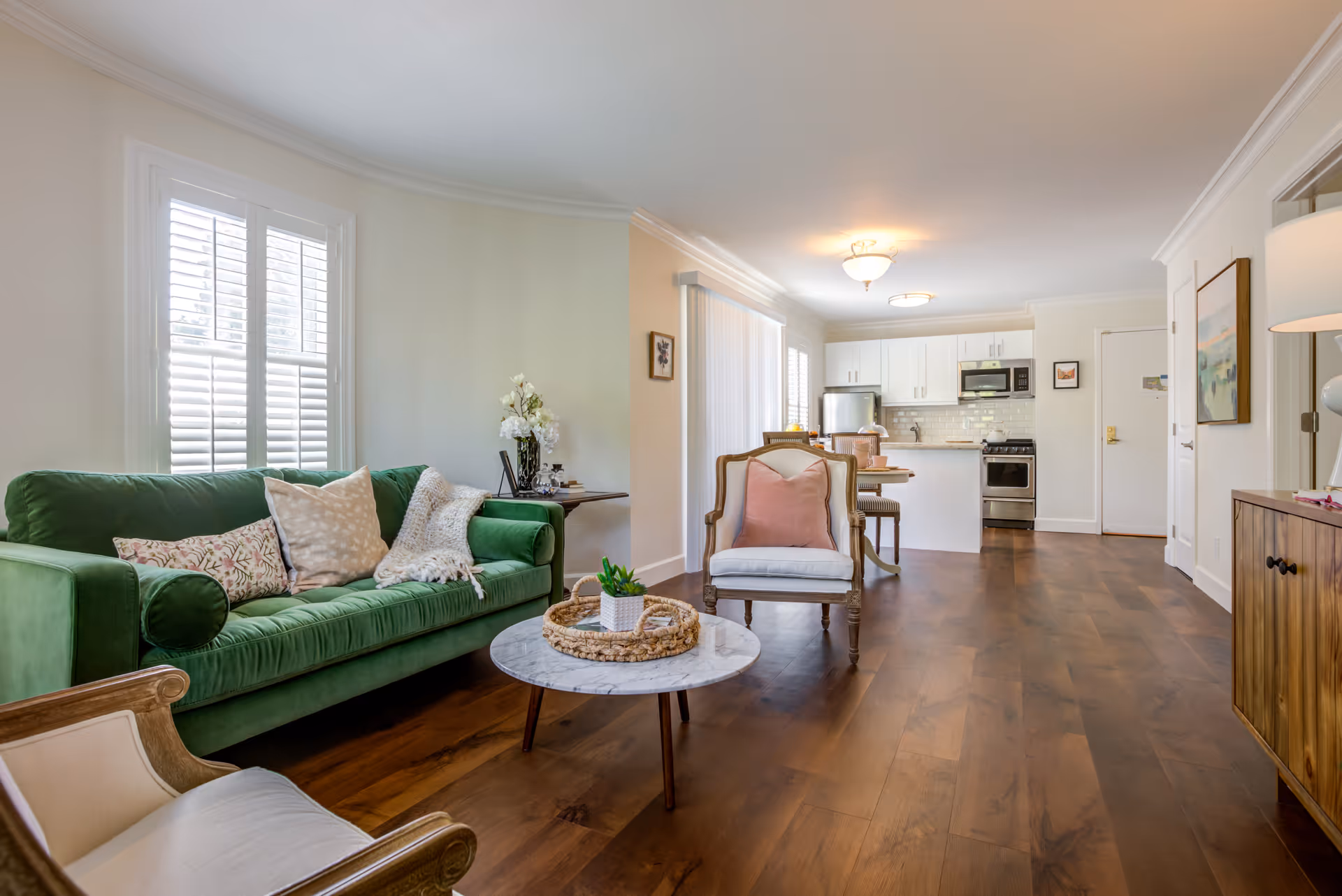 A bright and spacious living room and kitchen area with wooden flooring. The living room features a green sofa with decorative pillows, a round marble coffee table with a small plant, and two armchairs. The kitchen in the background has white cabinets, stainless steel appliances, and a small dining table with chairs. Large windows with white shutters allow natural light to fill the space.