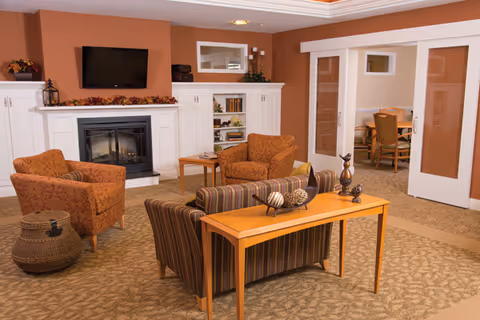 A cozy living room area with a striped sofa facing away from the camera, two patterned armchairs, a wooden console table with decorative items, a fireplace with a mounted flat-screen TV above it, built-in white shelves, and a doorway leading to a dining area with a table and chairs.