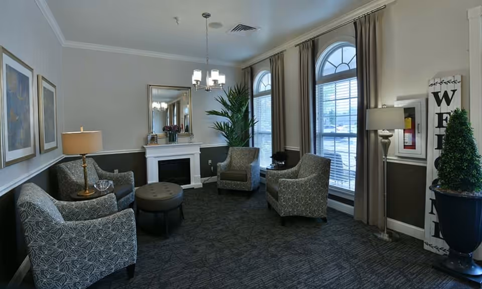 A cozy senior living facility lounge with four patterned armchairs arranged around a round ottoman. The room features a white fireplace with a mirror above it, two large arched windows with beige curtains, a tall potted plant, a floor lamp, and framed artwork on the walls. The carpet is dark with a subtle pattern, and the walls are painted in two tones with a chair rail.
