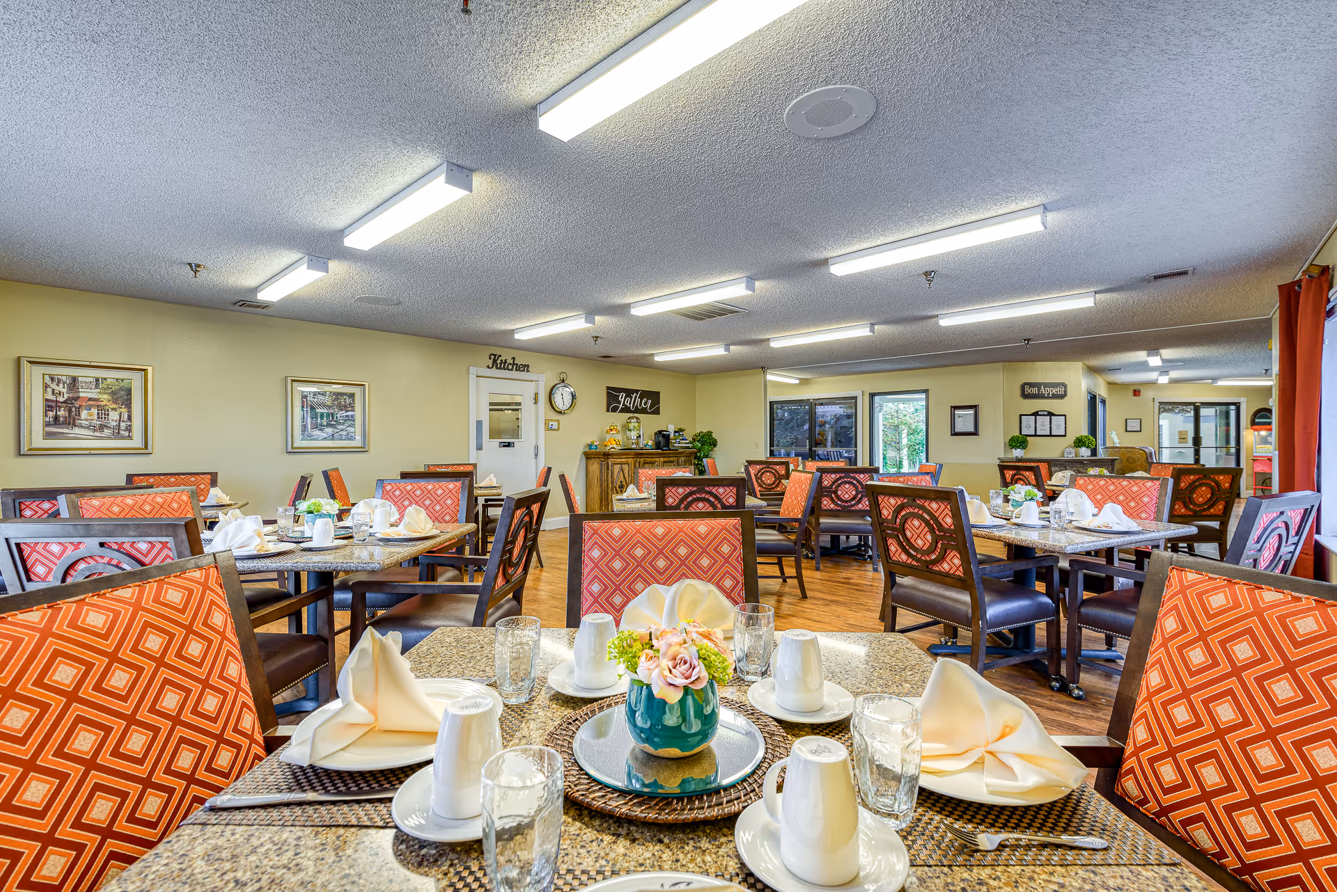 Communal dining room with tables set for meals, red patterned chairs, and a serving area in the back.
