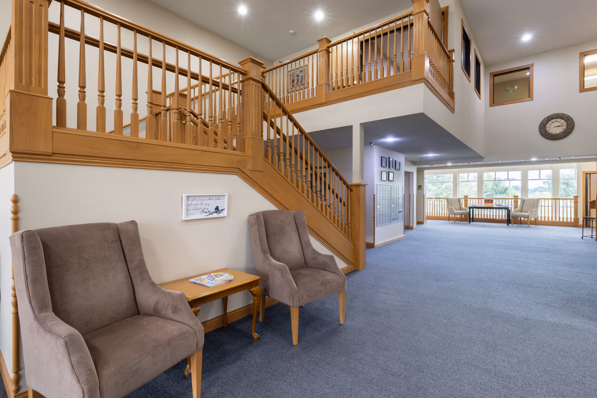 Interior view of a senior living facility showing a spacious common area with blue carpet, two gray upholstered chairs with a wooden side table between them, a wooden staircase with railings leading to an upper floor, and a seating area with chairs and a table near large windows letting in natural light.