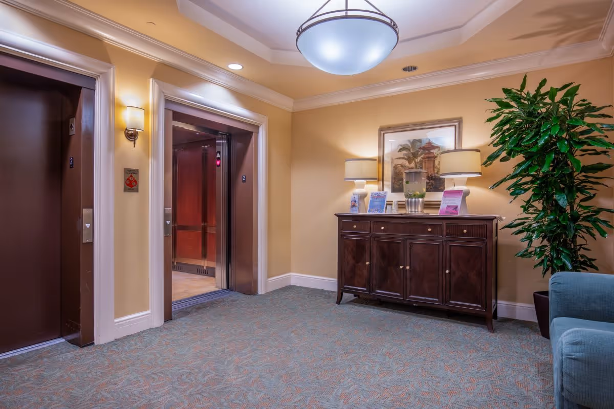 Interior view of a senior living facility hallway featuring two elevators, one with open doors. The area is warmly lit with beige walls, a patterned carpet, a wooden sideboard with two table lamps, informational pamphlets, a framed picture on the wall, and a large green potted plant next to a blue upholstered chair.