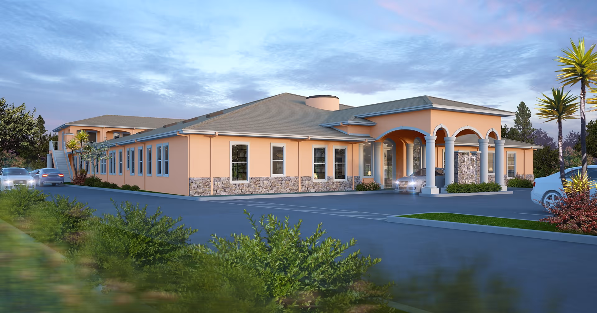 Exterior view of a single-story senior living facility building with peach-colored walls, stone accents, and a covered entrance supported by columns. The building is surrounded by a parking lot with several cars and landscaped greenery under a partly cloudy sky at dusk.