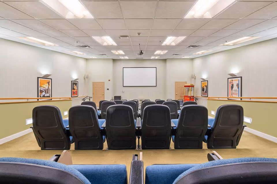 A small theater-style room with rows of blue and black cushioned chairs facing a blank projector screen on the front wall. The room has beige walls with framed posters, a popcorn machine in the corner, and ceiling lights.